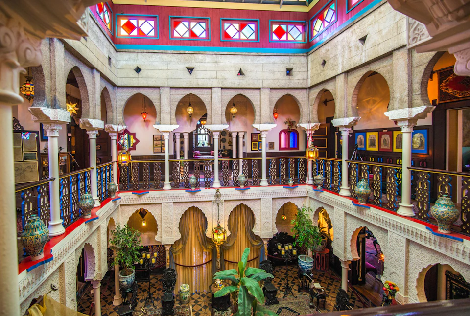 A vibrant, multi-story interior with archways, colorful stained glass, ornate railings, hanging lanterns, and potted plants.
