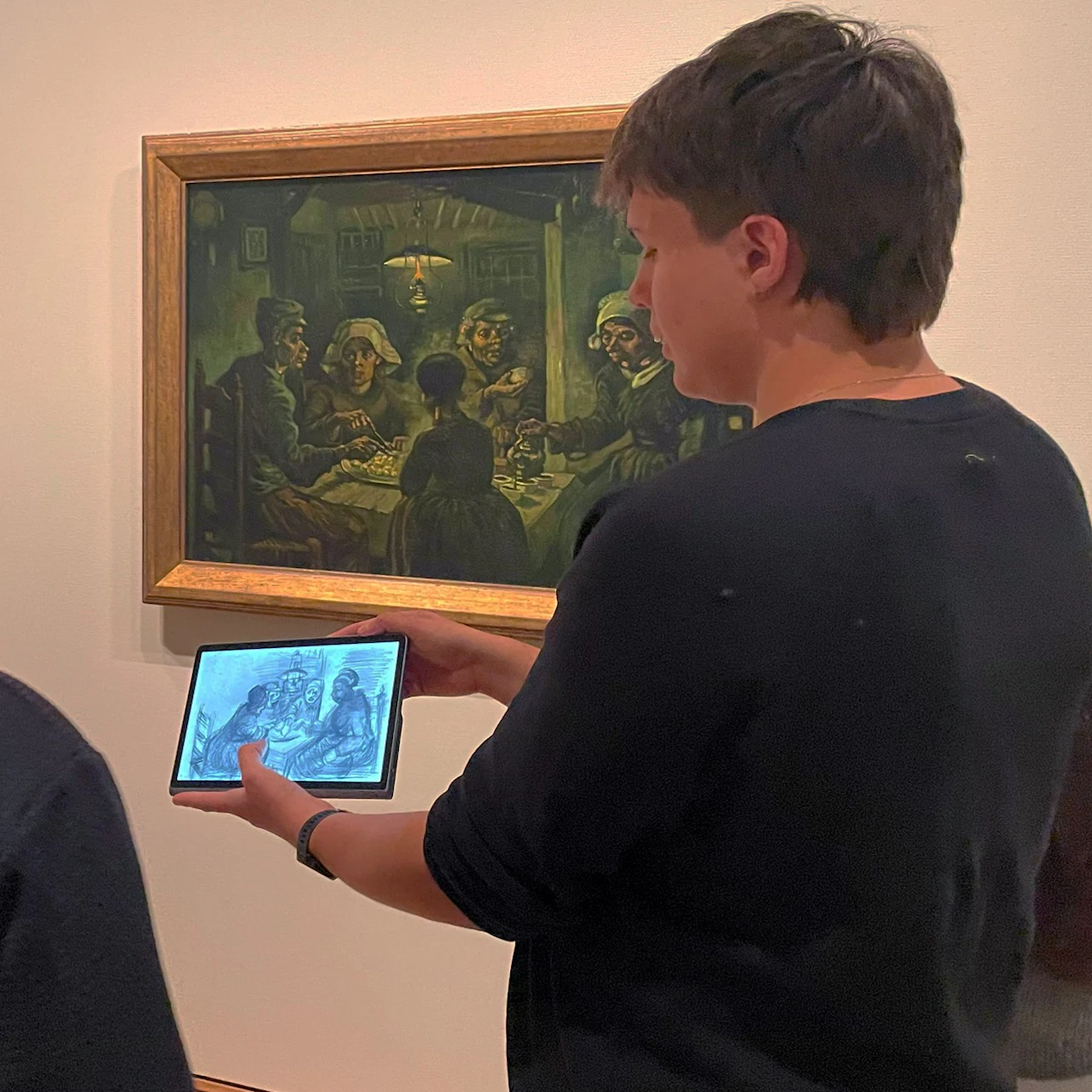 A person holding a tablet displaying an image in front of a framed painting on a museum wall.