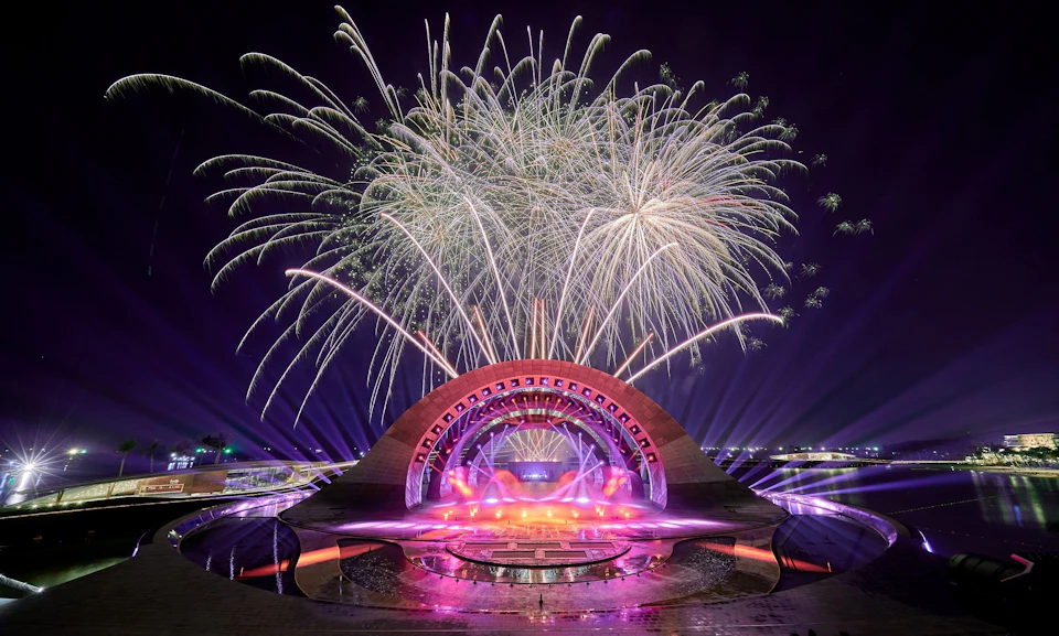 A vibrant fireworks display above a modern stage with colorful lights and an arched design, set at night.
