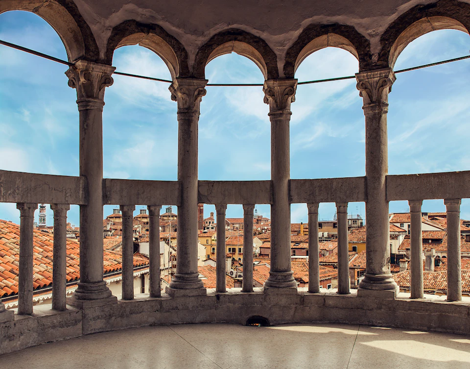 Balcony with arched columns overlooking a cityscape with red-roofed buildings under a partly cloudy blue sky.