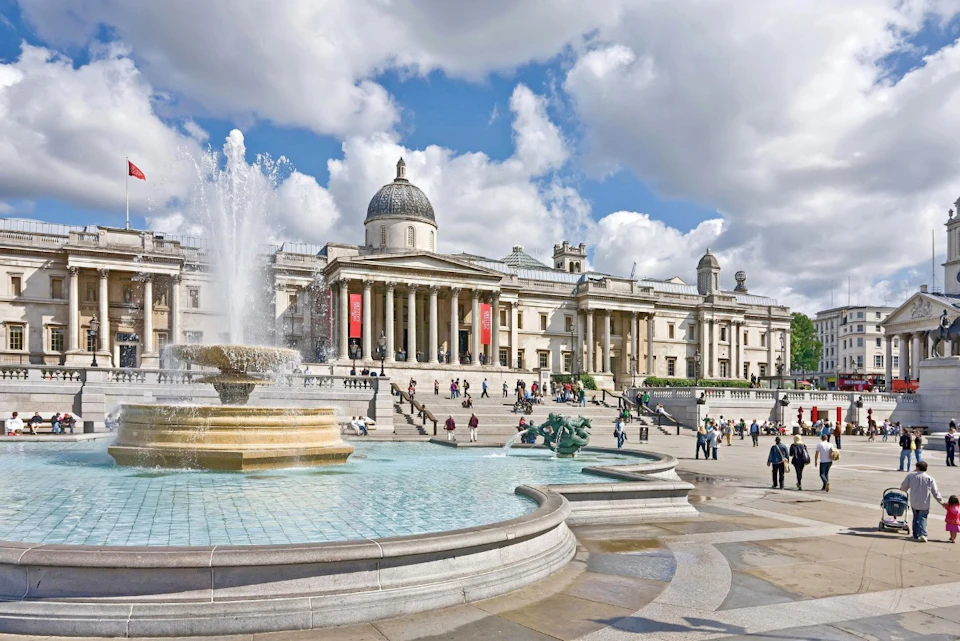 A grand neoclassical building with columns and a dome, surrounded by a square with fountains and visitors walking.