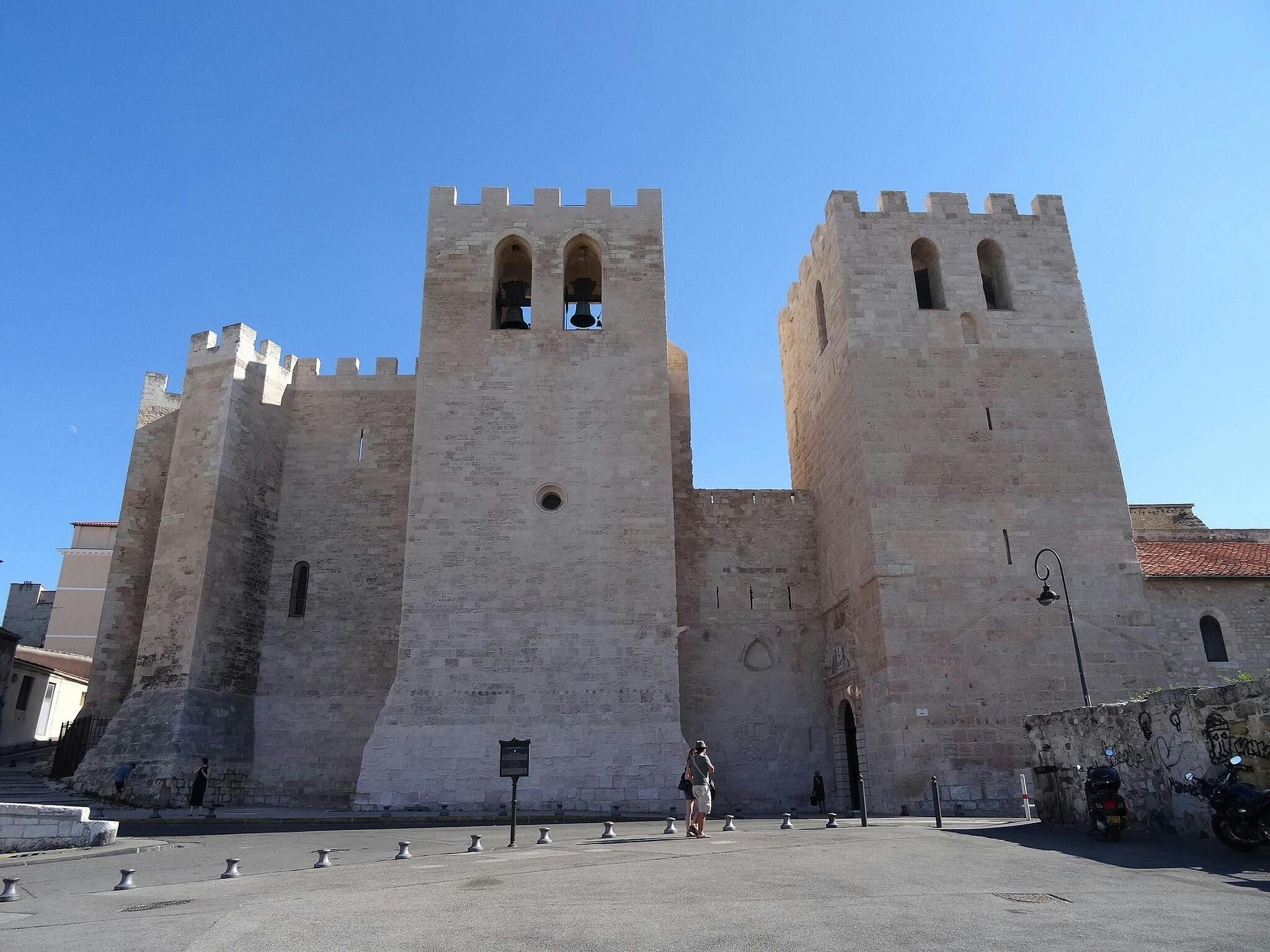 Три средневековые каменные башни с арочными окнами, несколько человек, идущих у входа, и ясное голубое небо.