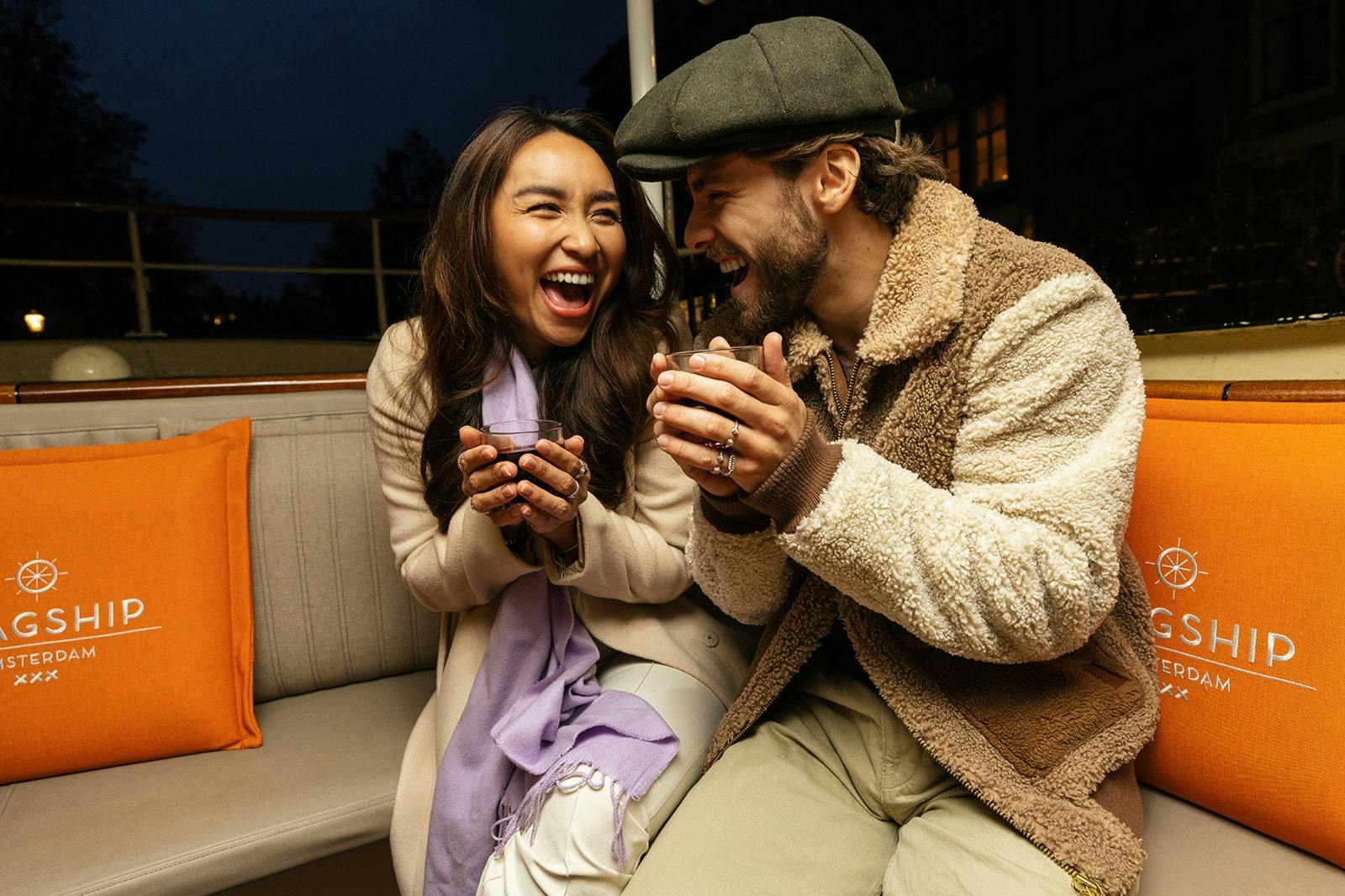 En kvinna och en man sitter på en bänk och ler och håller i muggar, med en orange kudde bredvid sig.