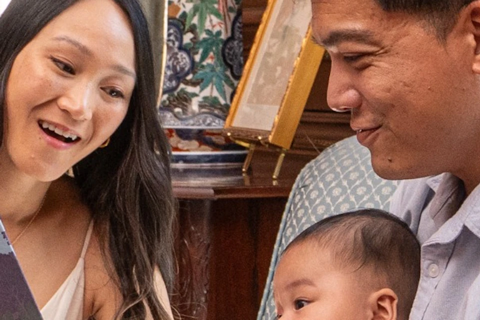 A couple sits on a chair in a warmly lit room, reading a book to a baby. The setting features wooden walls and large windows.