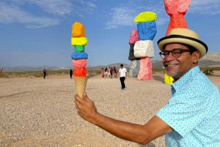 A man in a hat holds an ice cream cone, aligning it with colorful stacked rocks in a desert landscape.