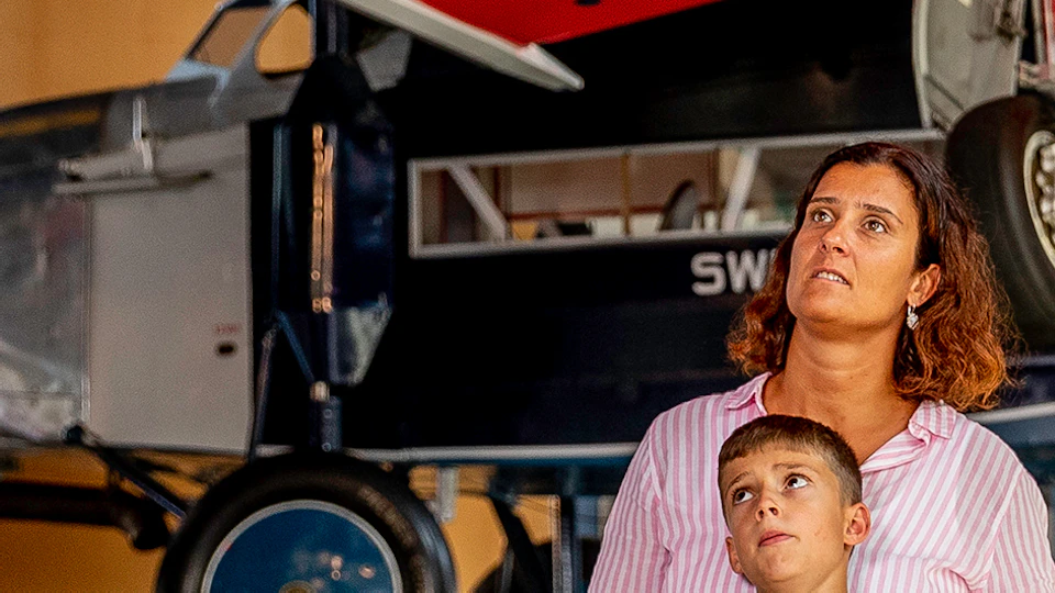A family of four looks up at a red airplane displayed in a museum, with the child on the father's shoulders pointing at the plane.
