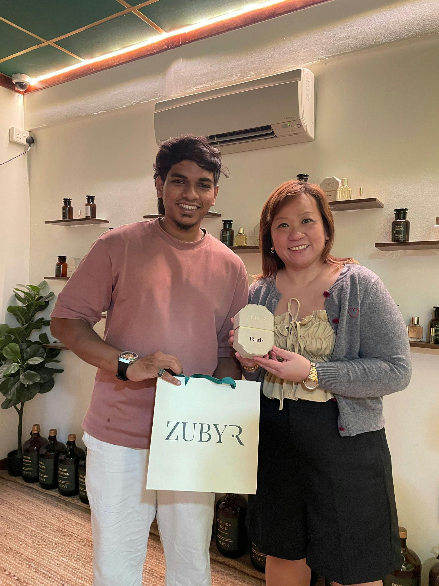 Two people smiling indoors, holding a gift bag and a wooden item with the name "Ruth" engraved on it. Shelves with bottles are in the background.