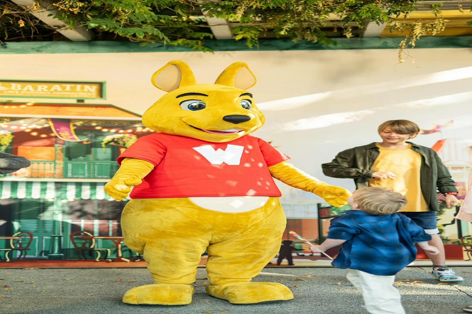 A person in a yellow kangaroo costume with a red shirt stands on stage near a child and an adult under a leafy canopy.