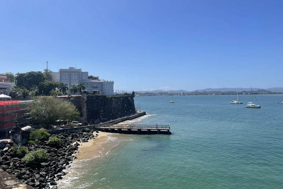 A coastal scene with a white building on a cliff, a small sandy beach, a pier, and calm blue water under a clear sky.
