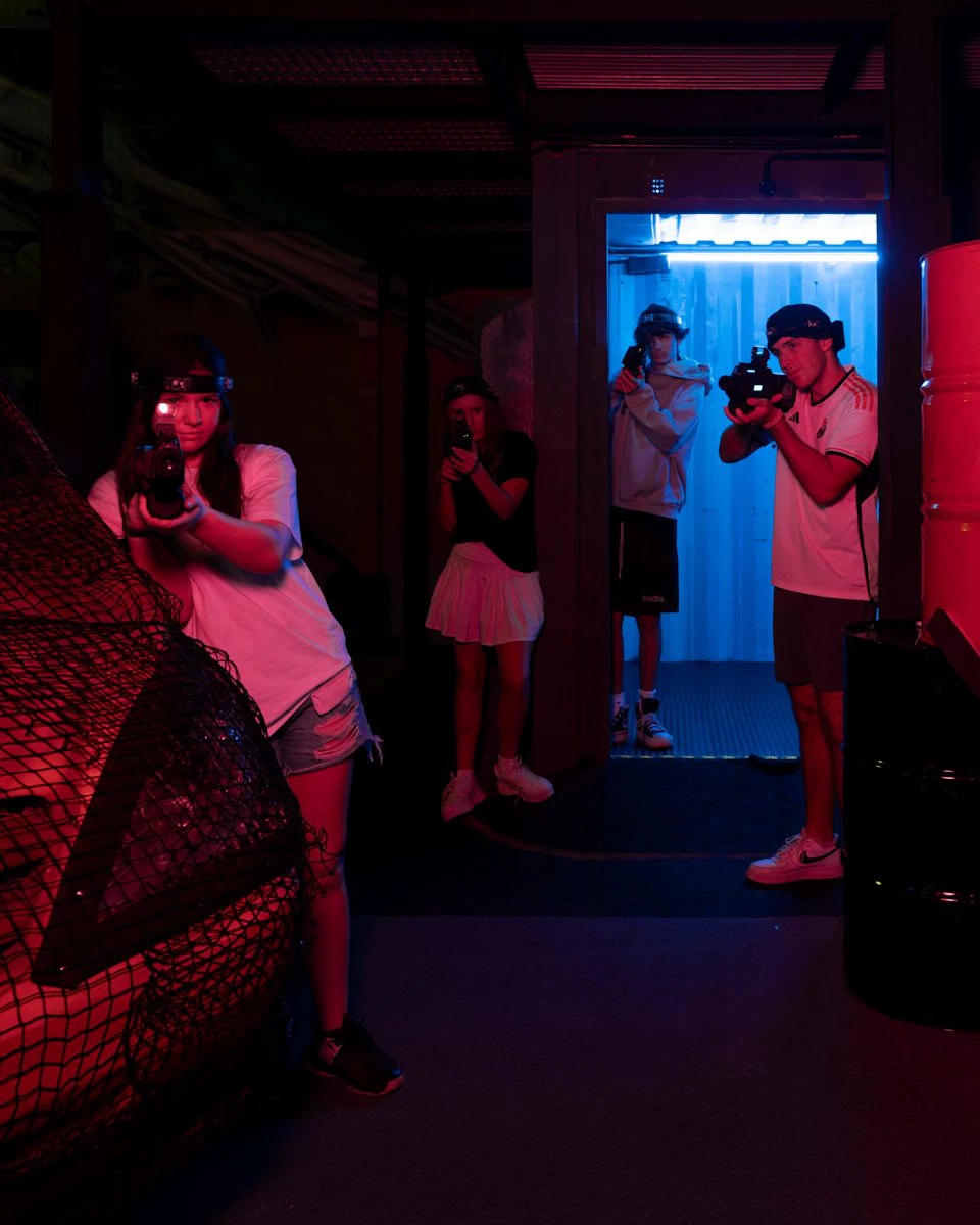 Four people are playing laser tag in a dimly lit arena with red and blue lighting, holding laser guns and wearing headbands.