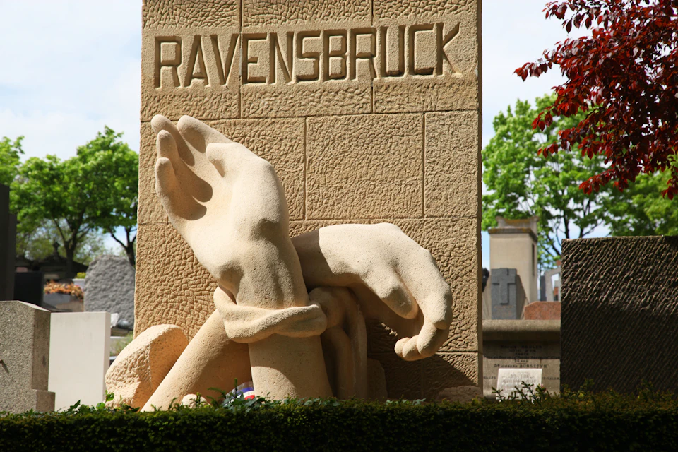A stone monument displays the word "RAVENSBRUCK" with two large sculpted hands, one in chains, against a backdrop of trees and gravestones.