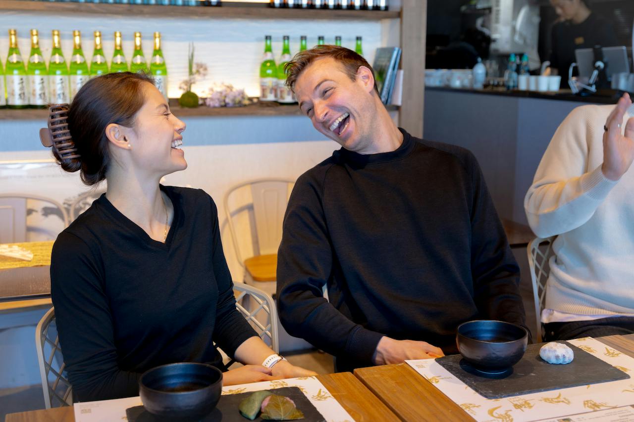 Two people sitting at a table in a restaurant, smiling and laughing with each other. Bottles are visible in the background.