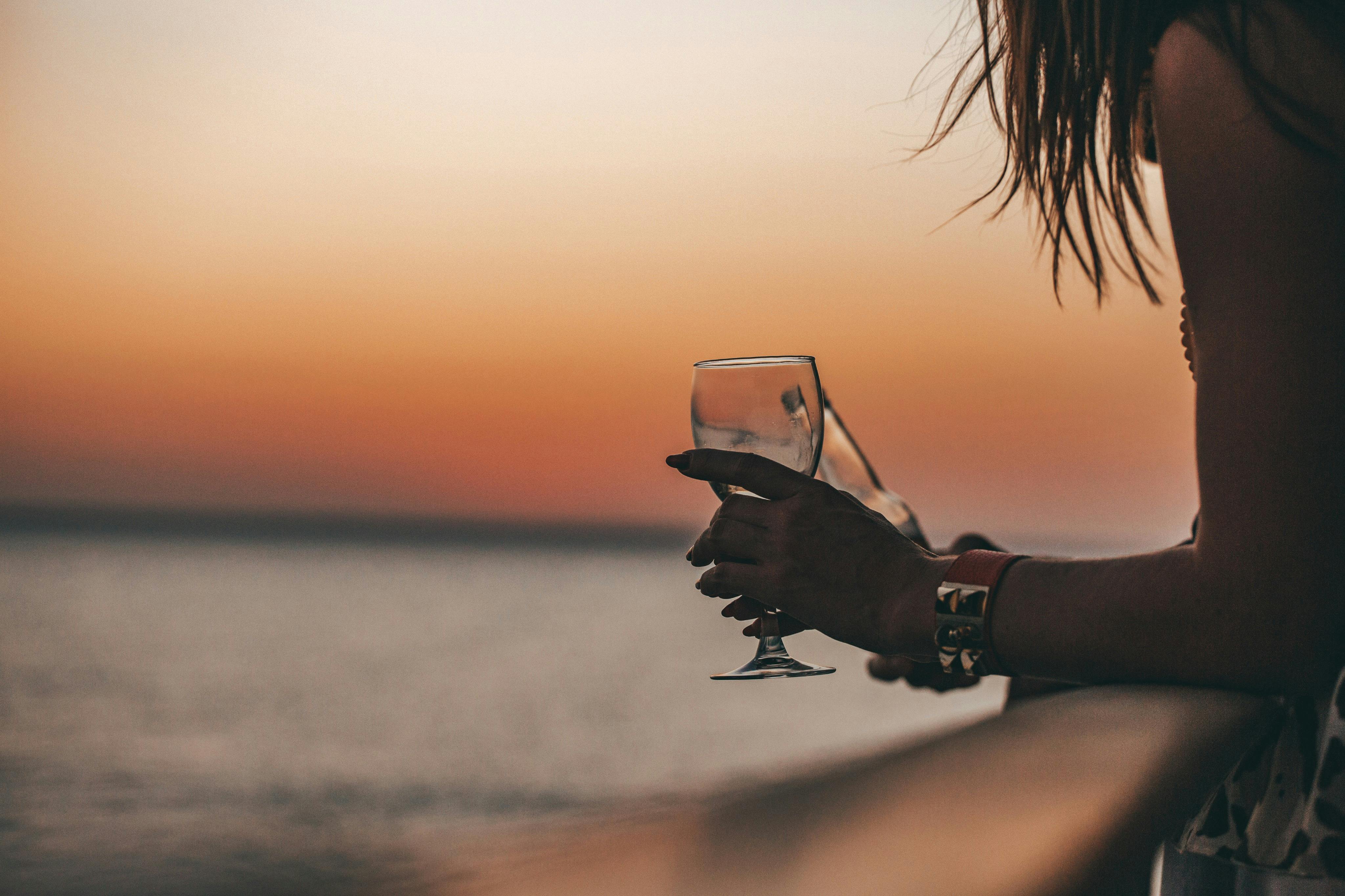 A person holding a wine glass by a railing, overlooking a calm sea at sunset with an orange and pink sky.