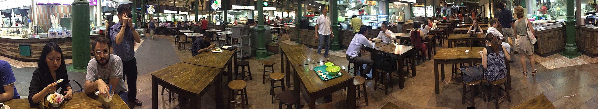 Mensen dineren en praten aan houten tafels in een drukke foodcourt, met verschillende eetkraampjes op de achtergrond.