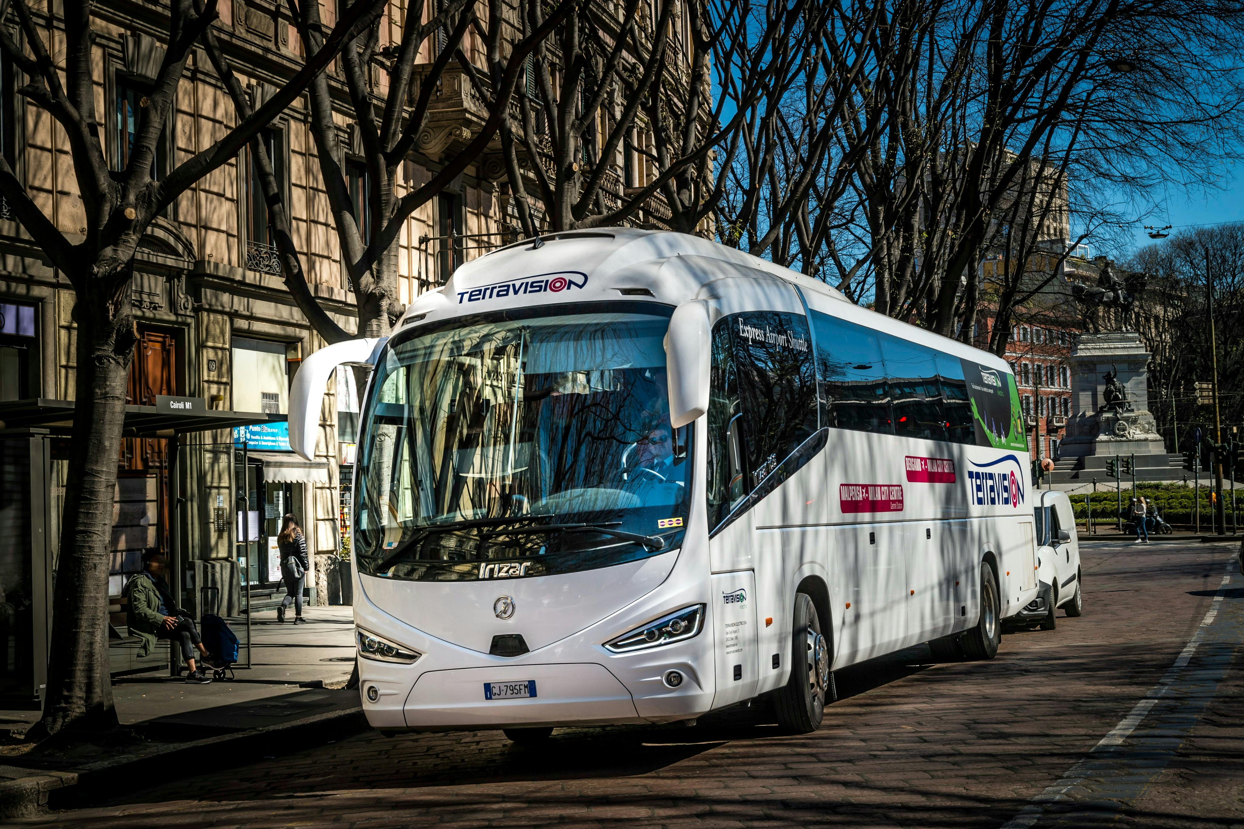 Ein weißer Terravision-Bus parkt auf einer von Bäumen gesäumten Straße mit Gebäuden und einer Bushaltestelle im Hintergrund.