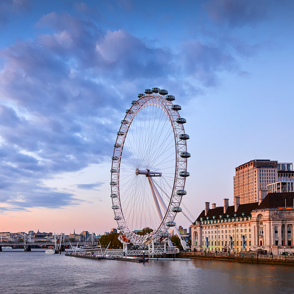 Large Ferris wheel next to a river, with buildings and a bridge in the background under a partly cloudy sky.