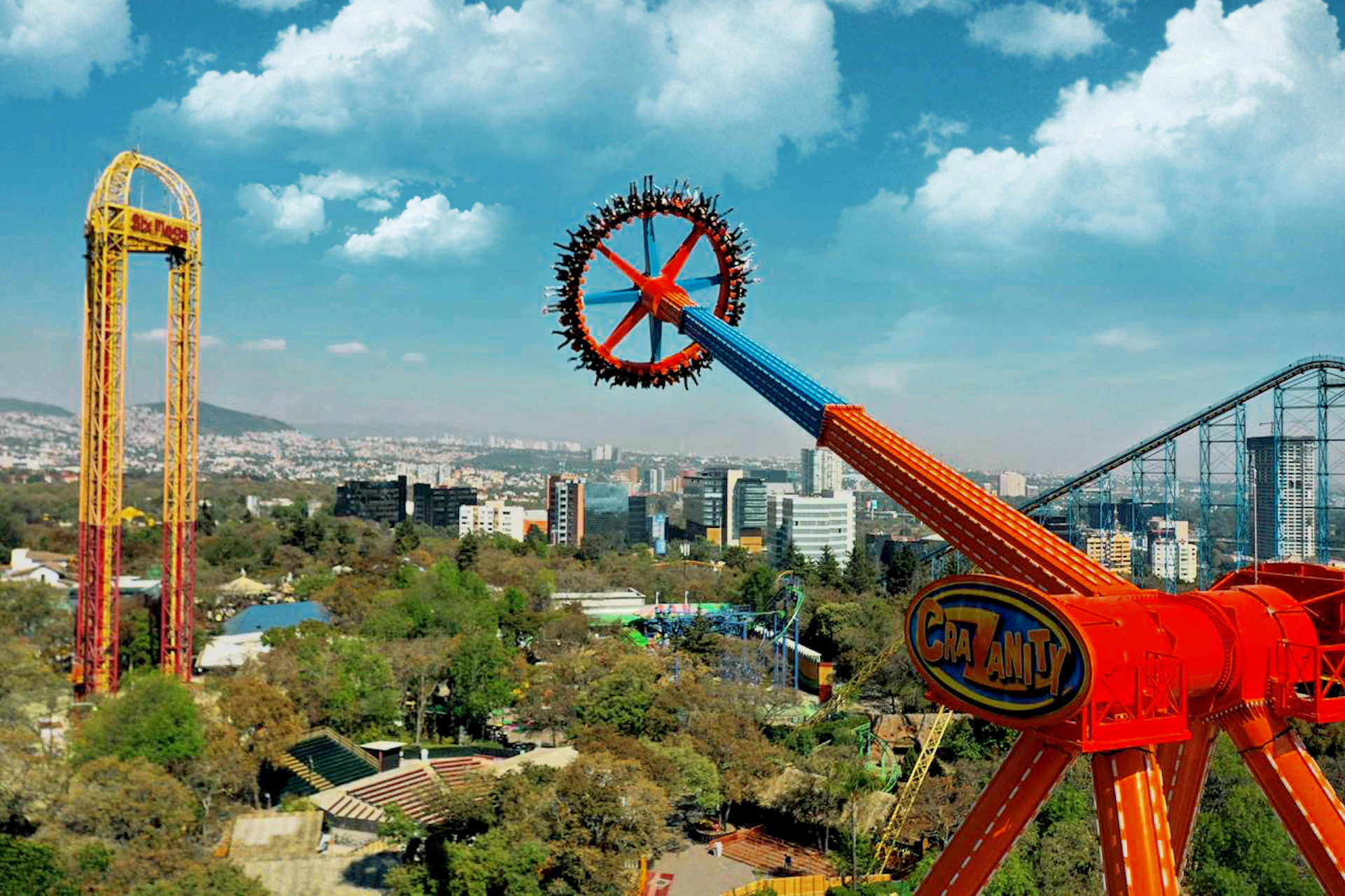 Amusement park ride in motion, spinning high above a cityscape with buildings, trees, and a blue sky with clouds in the background.
