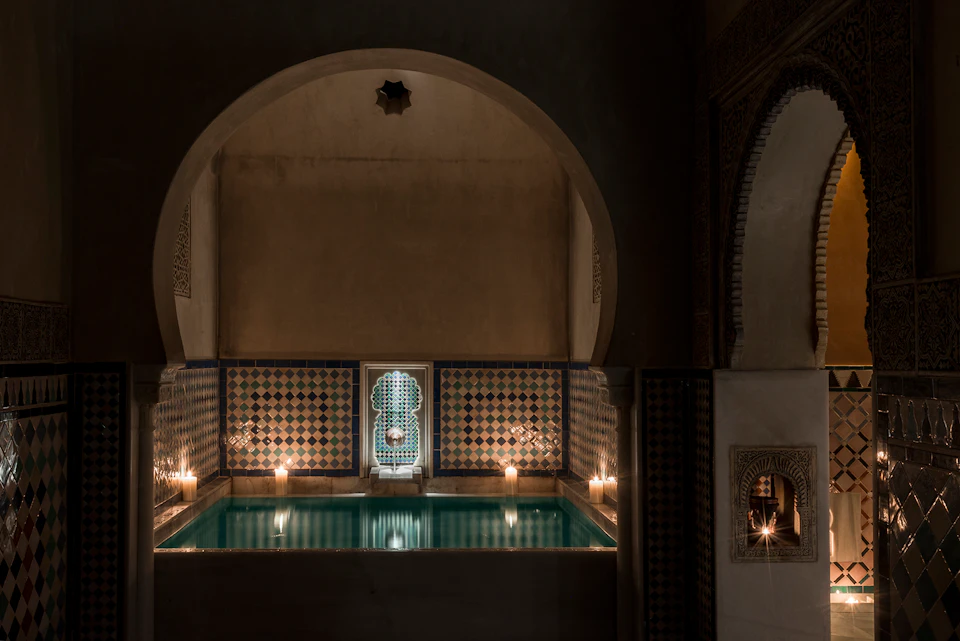 A dimly lit bathhouse features an ornate tiled pool, candles, archways, and intricate wall detailing.