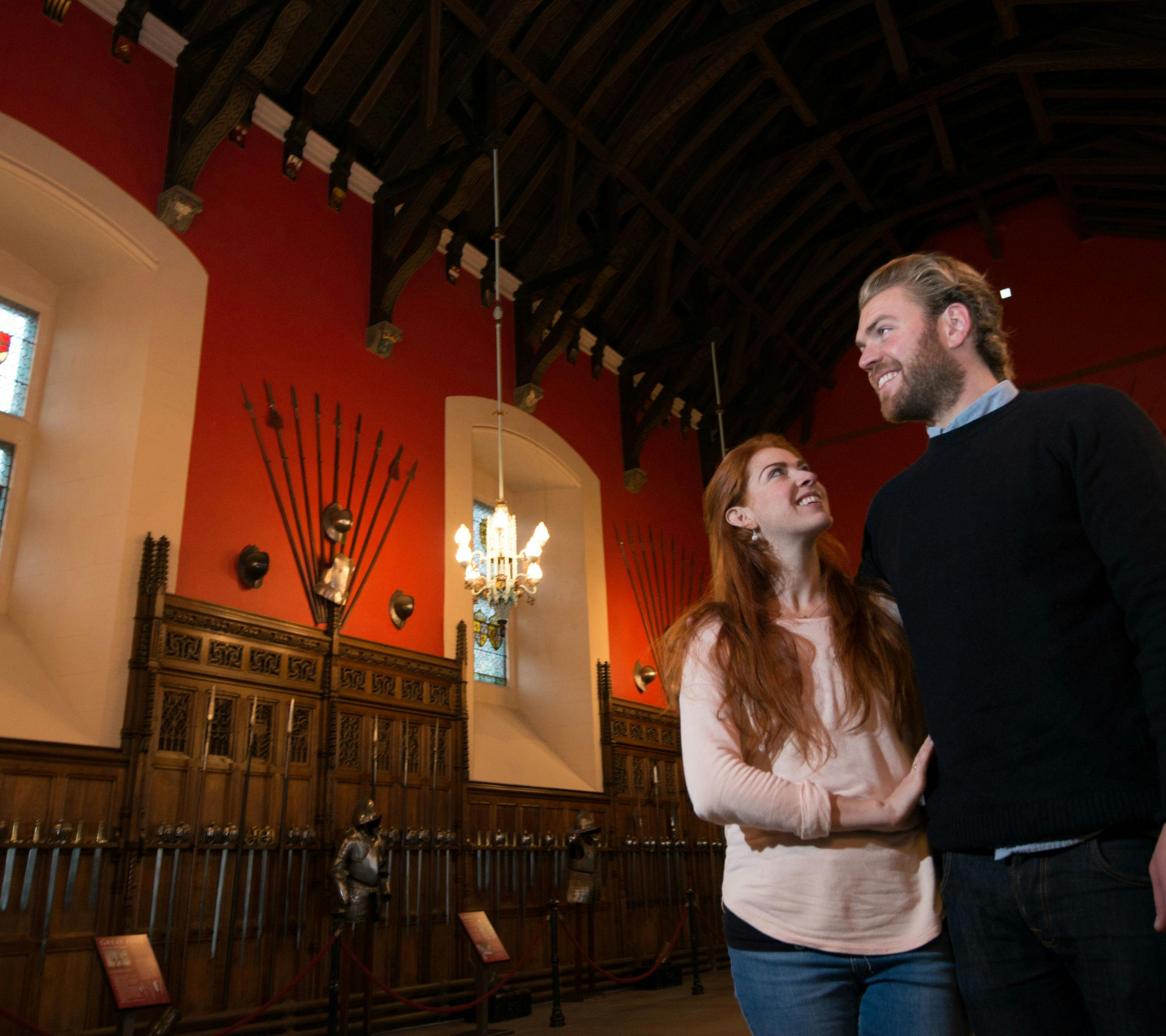 Due persone si trovano in una sala del museo poco illuminata con pareti rosse, armature medievali e un lampadario che pende da un soffitto alto.