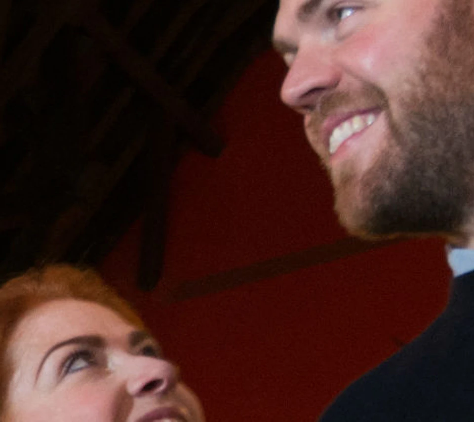 Two people stand in a dimly lit museum room with red walls, medieval armor displays, and a chandelier hanging from a high ceiling.