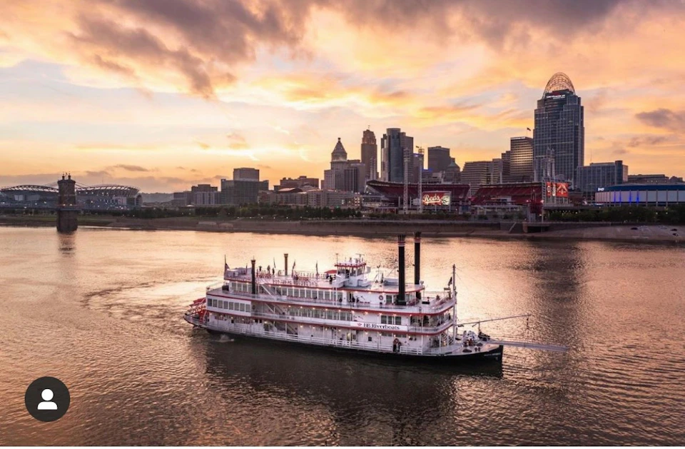 A riverboat cruises on a river at sunset with a city skyline, including buildings and a stadium, visible in the background.