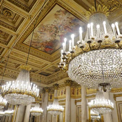 Utsmyckat rum med stora kristallkronor, pelare och en detaljerad takmålning som visar en klassisk scen.