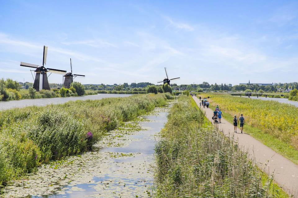 A scenic path beside a canal with water plants, flanked by windmills and green fields, with people walking under a blue sky.