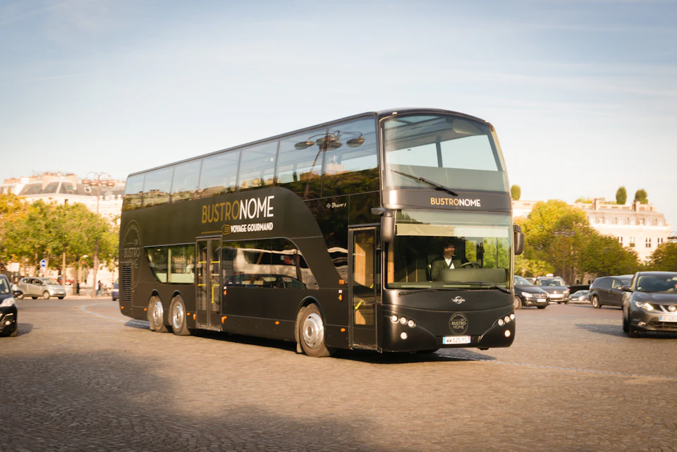 Black double-decker bus labeled "BUSTRONOME" and "VOYAGE GOURMAND" driving on a cobblestone road with trees in the background.