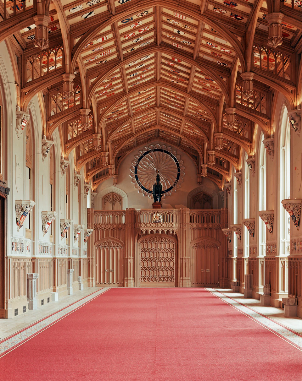A grand hallway with an ornate wooden ceiling, arched windows, red carpet, and a large decorative wheel at the far end.