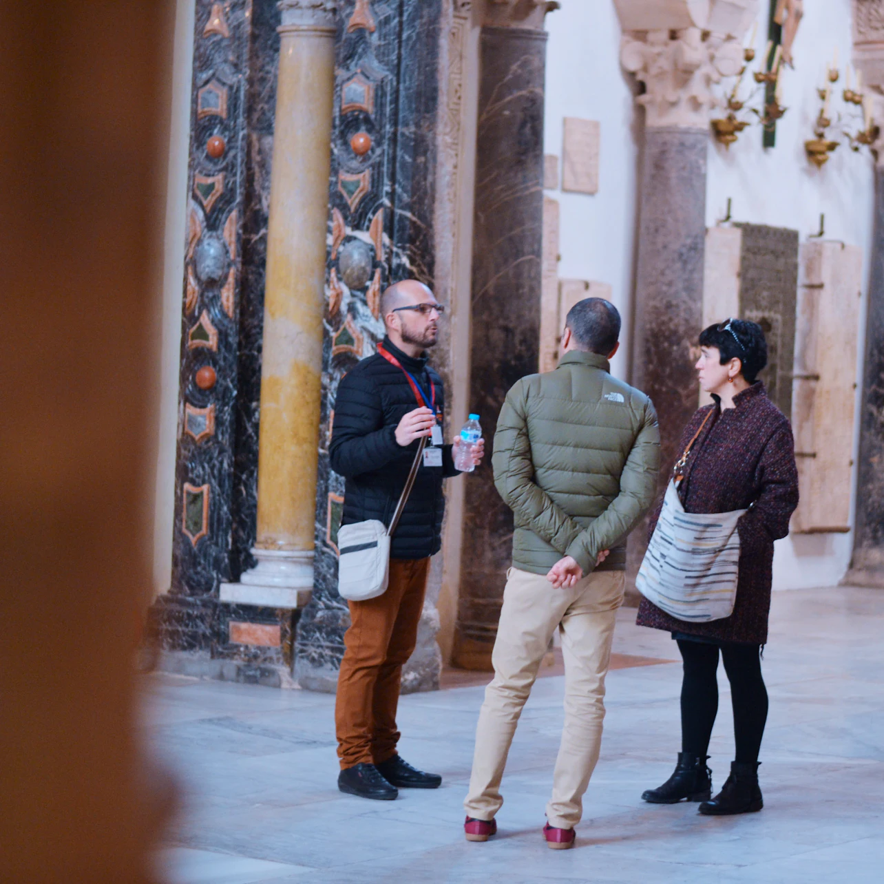Three people stand talking in a historical building with ornate marble columns and stone decorations in the background.