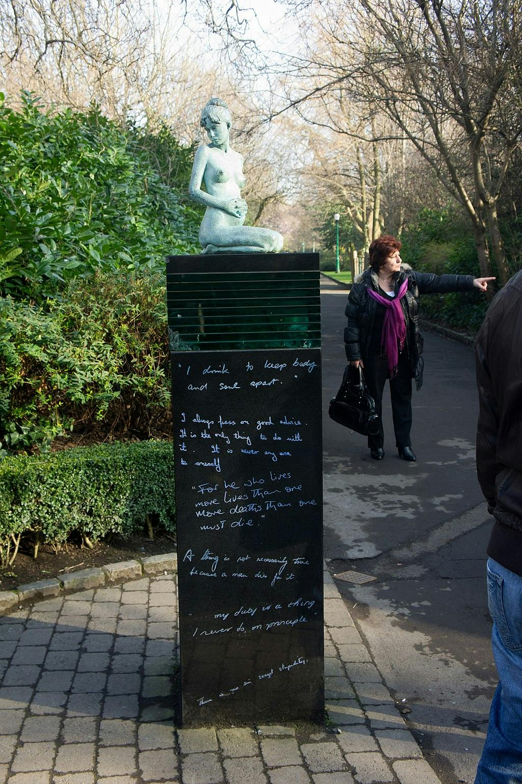Bronzová socha klečící ženy na podstavci s nápisem v parku. Osoba ukazuje směr na nedaleké cestě.
