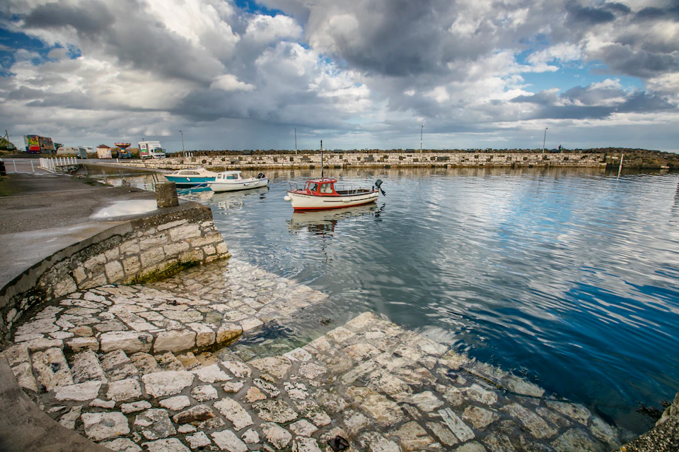 Stone-paved dock with two small boats anchored on calm water under a partly cloudy sky.