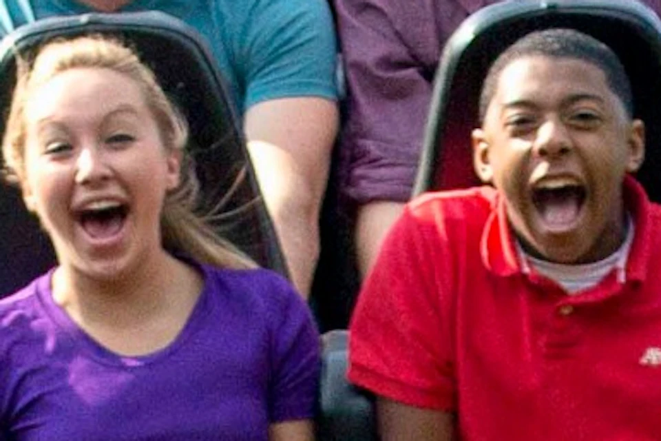 A group of people on a wooden roller coaster, smiling and raising their hands, riding in a red train labeled "The Beast."