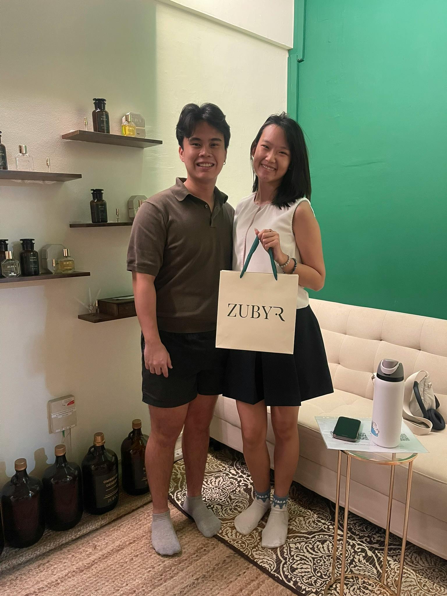 Two people standing and smiling indoors, with the person on the right holding a shopping bag labeled 'ZUBYR.' Shelves with bottles and a green wall are visible in the background.