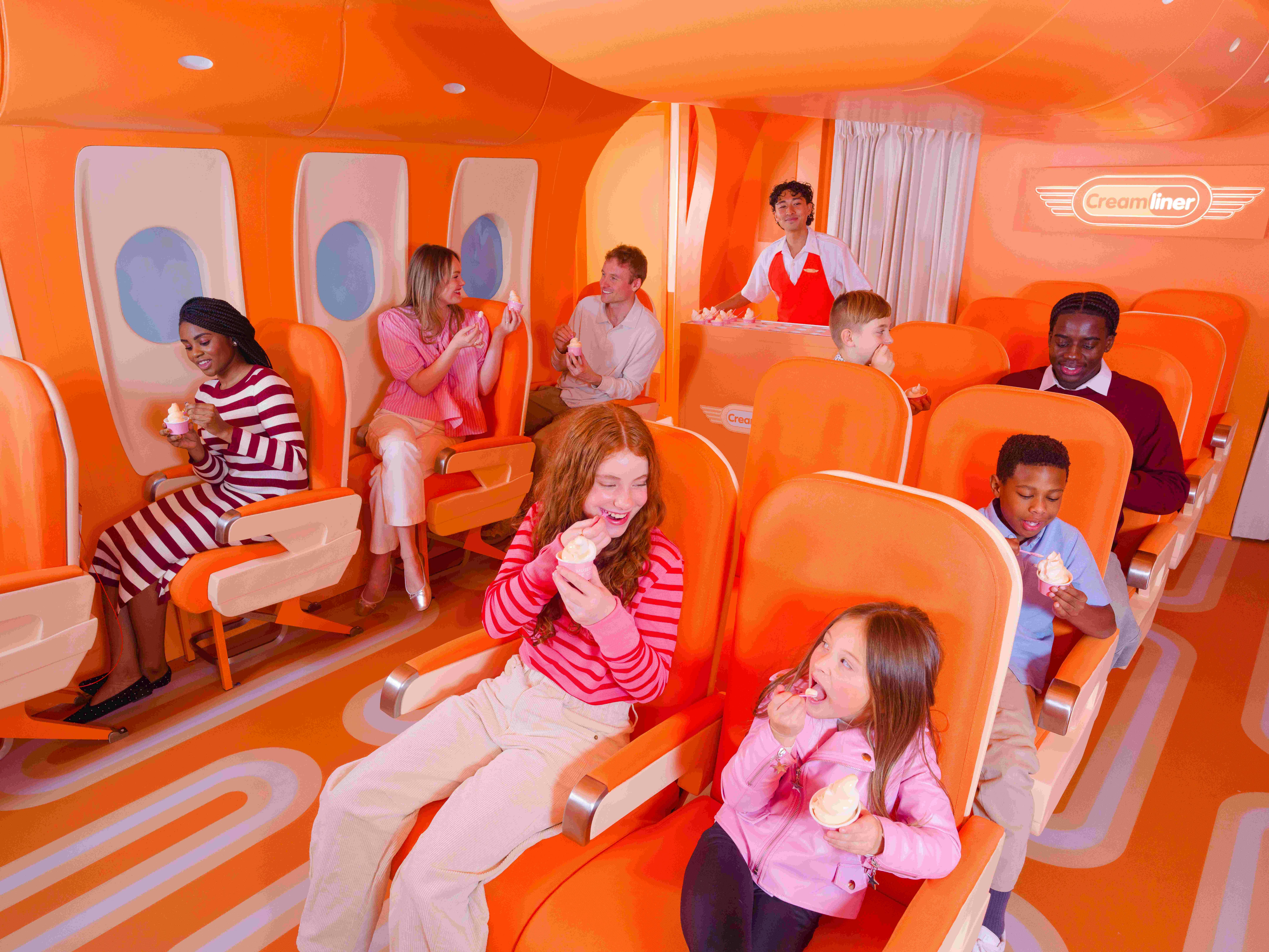 People, including children, enjoying ice cream inside a brightly colored, orange airplane-themed room with seat rows and windows.