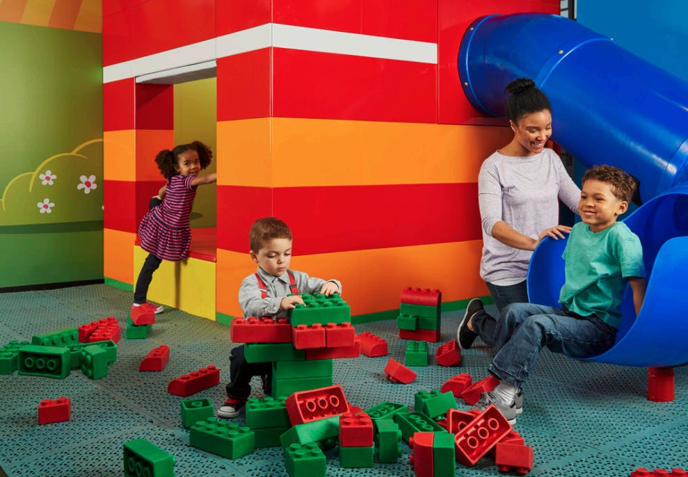 Children playing with large, red and green building blocks beside a colorful play structure with a blue slide. An adult supervises.