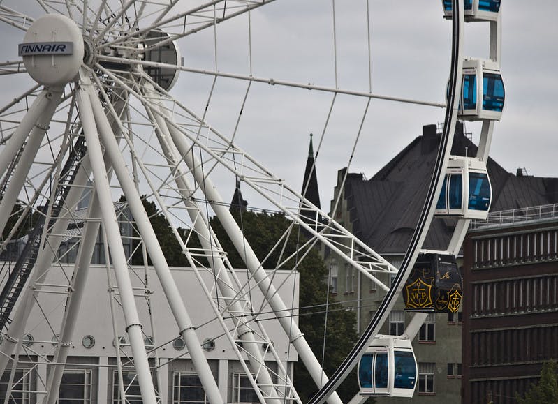 Close-up of a Ferris wheel with blue cabins, set against an urban backdrop featuring buildings and trees under a cloudy sky.