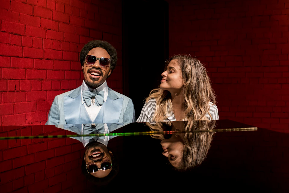 A man in a blue suit and sunglasses sits at a piano with a woman in a striped shirt, both in front of a red brick wall.