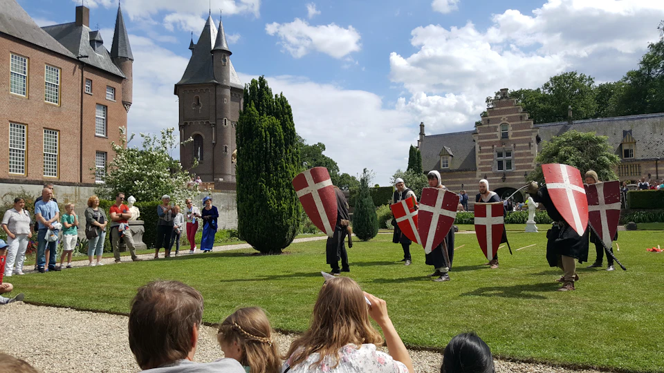 Performers in medieval costumes holding shields with crosses, standing on a lawn. A castle-like building is in the background.