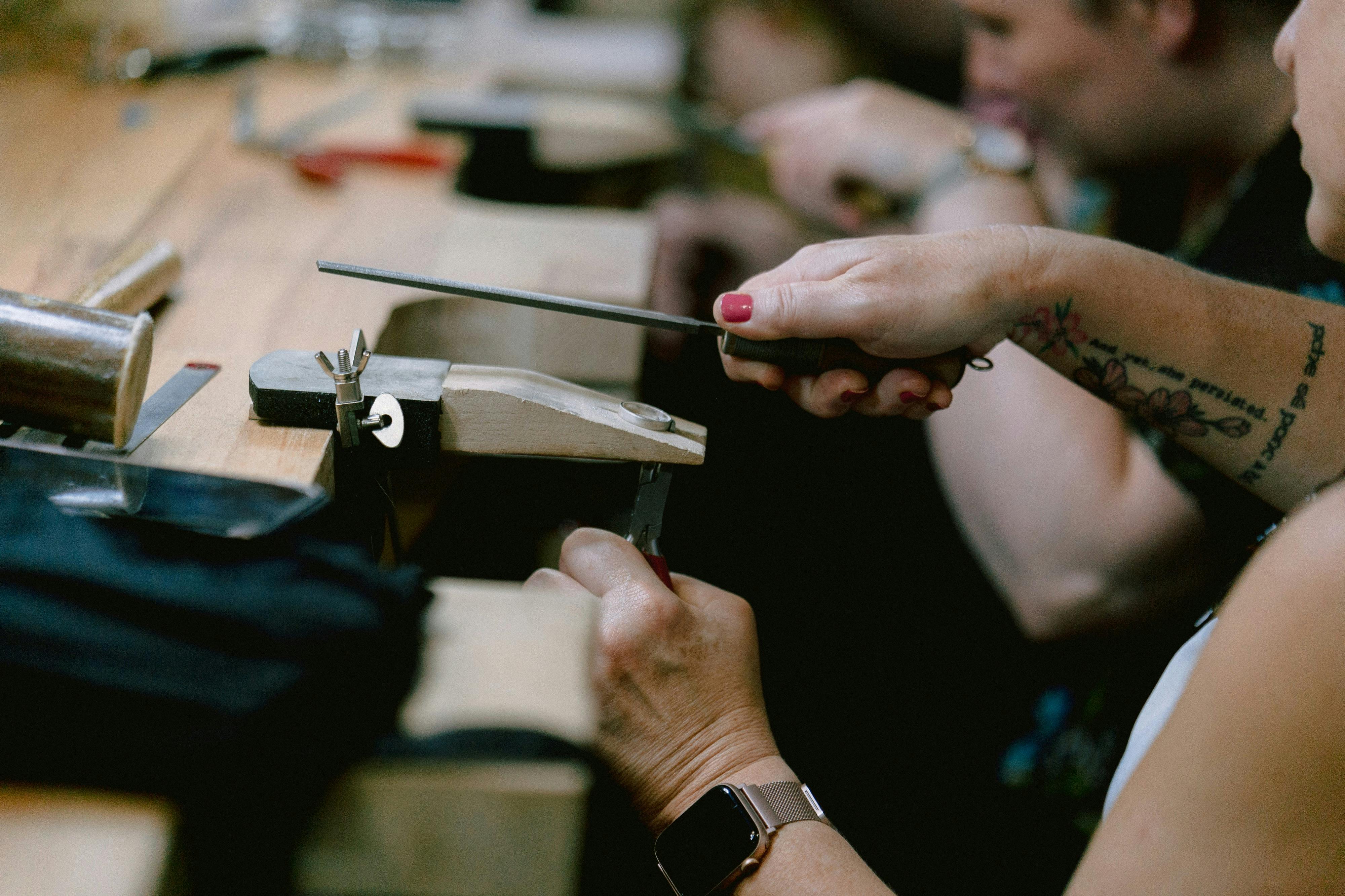 People working with hand tools at a bench, focusing on a person's tattooed arm and wristwatch.