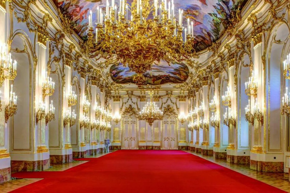 An ornate hall with golden chandeliers, decorated ceiling, and walls lined with mirrors, featuring a red carpet floor.