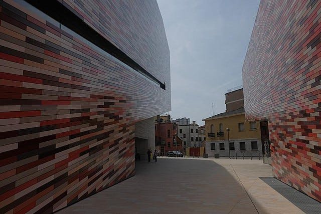 Moderna byggnader med flerfärgade rektangulära plattor skapar en smal passage som öppnar till ett litet torg med andra byggnader.