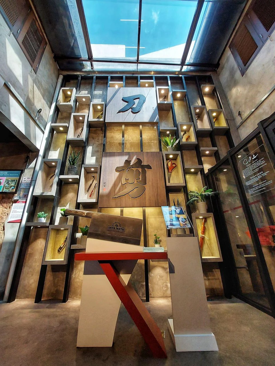 A modern indoor display wall with wooden shelves showcasing knives and decor items, centered around a large character plaque.