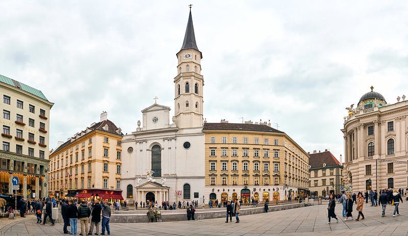 높은 첨탑이 있는 유서 깊은 교회는 흐린 날 여러 개의 베이지색 건물로 둘러싸여 있습니다. 사람들은 걷고 전경에 모입니다.