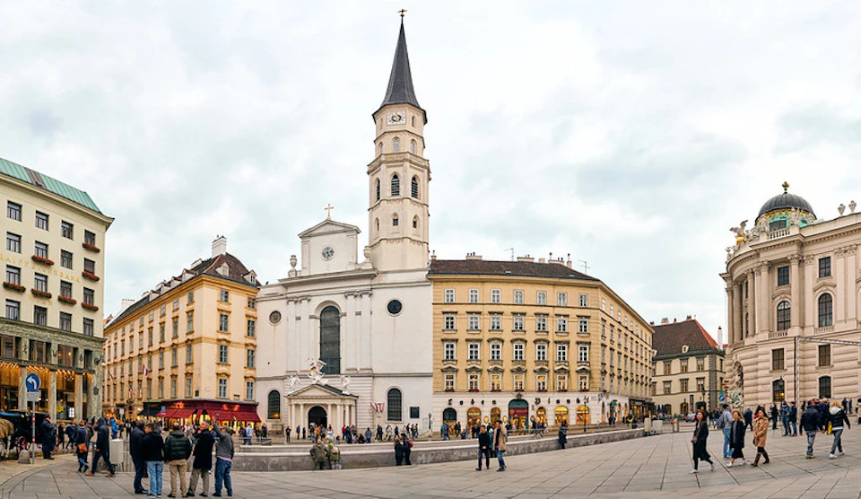 A historic church with a tall steeple is surrounded by several beige buildings on a cloudy day. People walk and gather in the foreground.