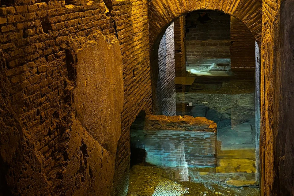 A dimly lit underground corridor with brick walls, arches, and a reflective water surface on the floor.