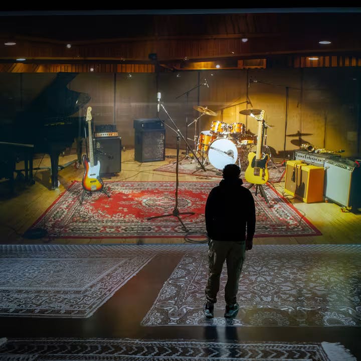 Osoba stojí před sestavou nahrávacího studia s kytarami, bicí soupravou, mikrofony a zesilovači na červeném koberci.