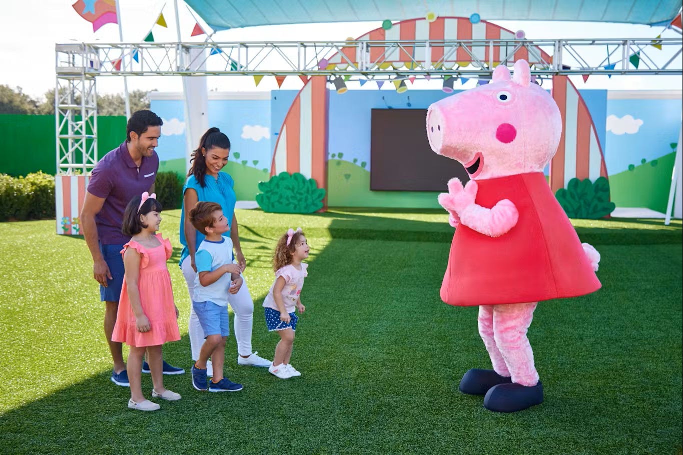 A family with two children interacts with a person in a pig costume in a colorful outdoor play area.