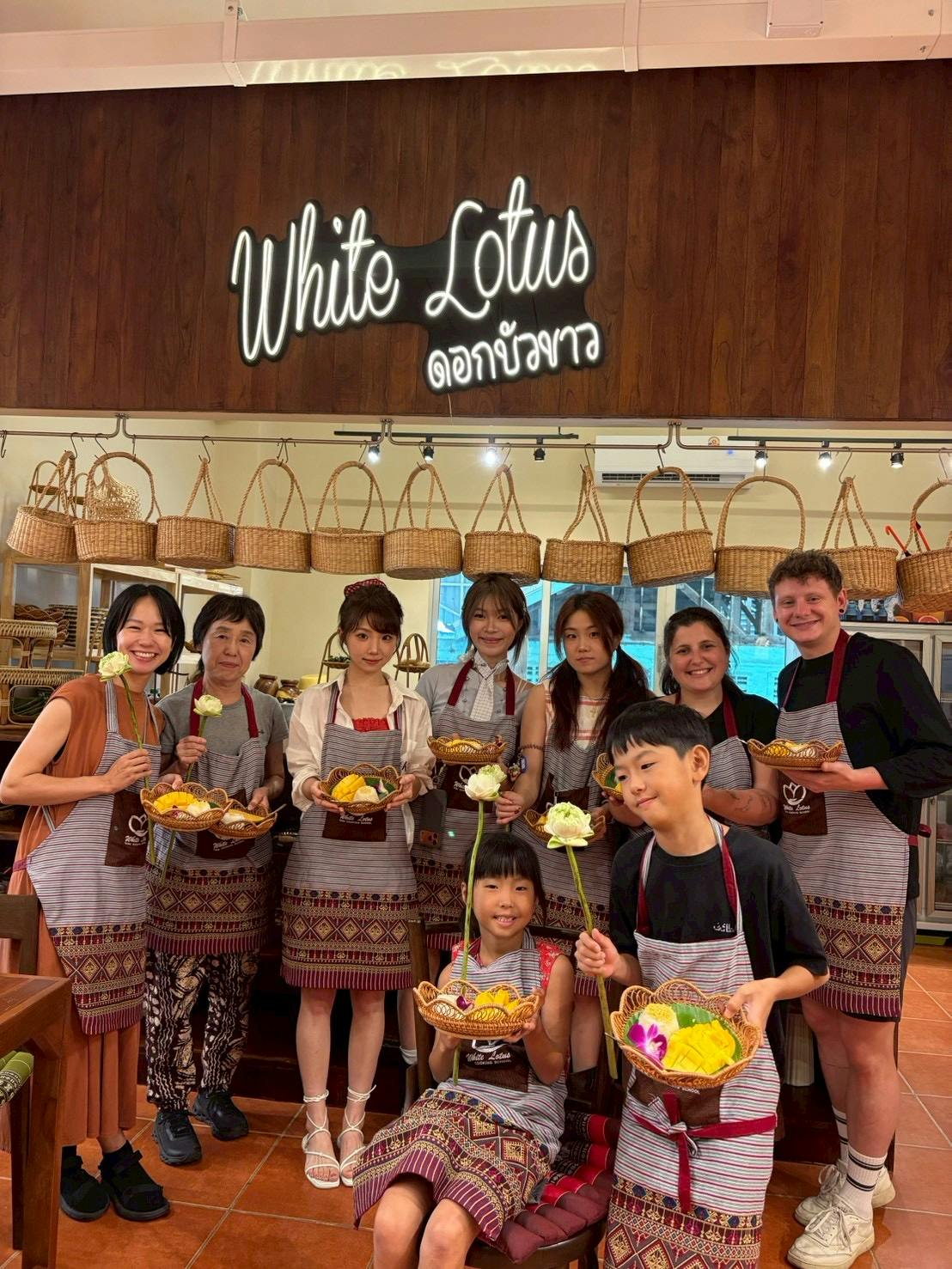 A group of people posing indoors wearing aprons, holding food baskets, and smiling. A sign and baskets are visible behind them.