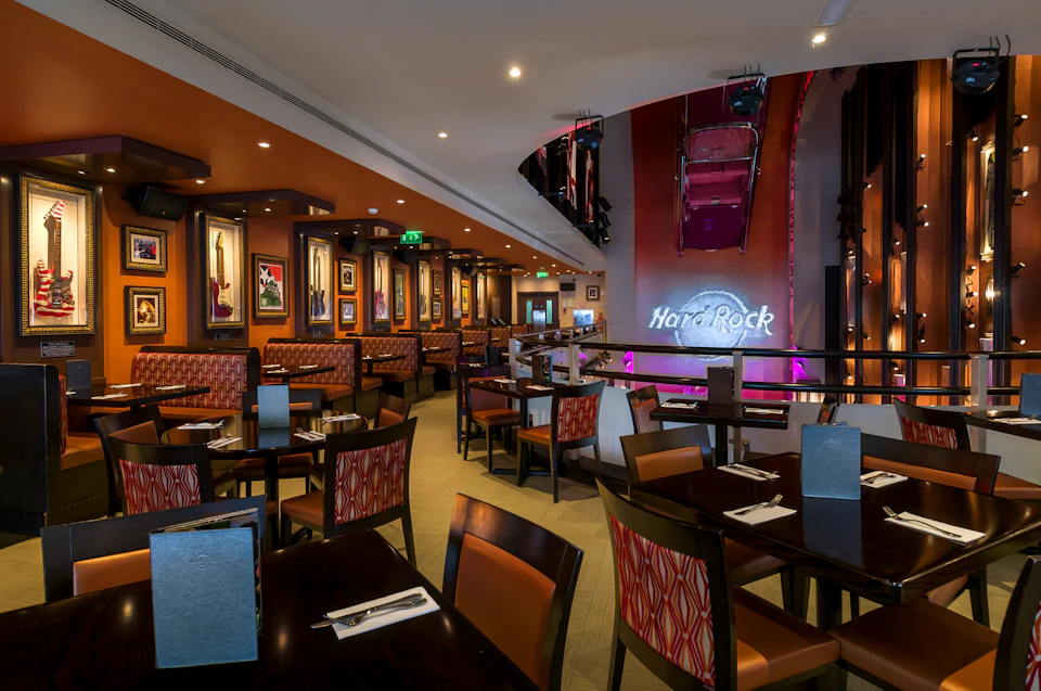 Modern restaurant interior featuring wooden tables and chairs, vibrant framed memorabilia, and a lit "Hard Rock" sign on the wall.